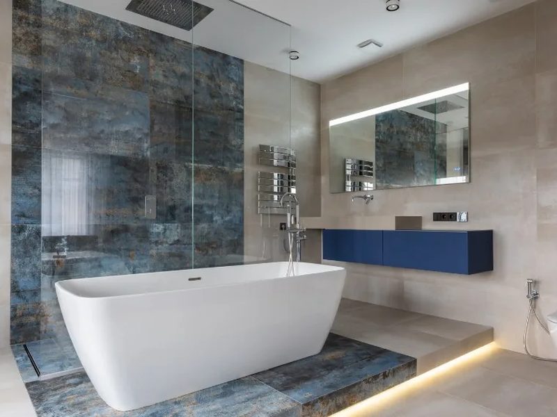 Baño con bañera exenta sobre tarima iluminada y revestimiento efecto piedra azul