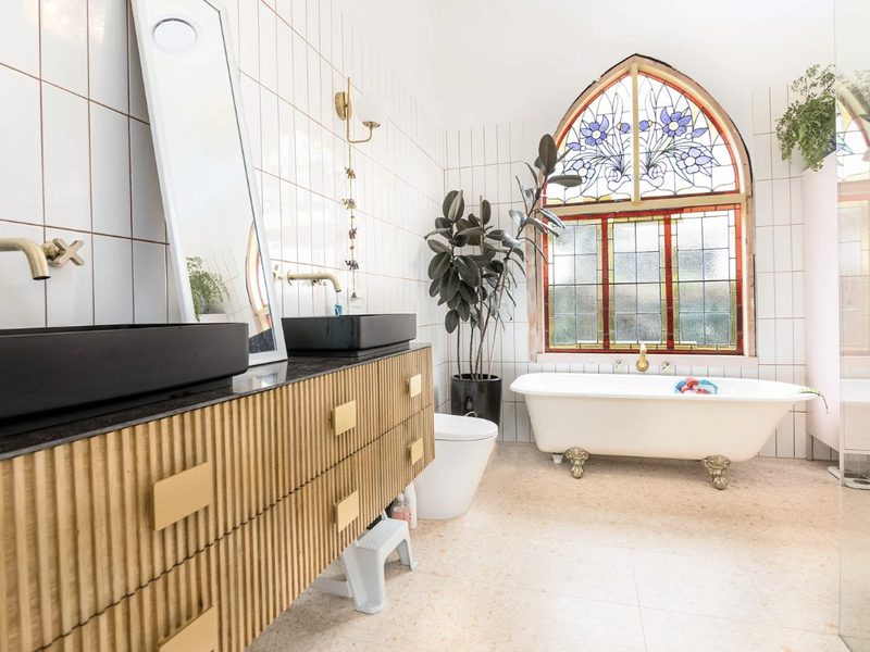 Baño reformado con mueble de madera acanalada, doble lavabo negro, bañera de patas y ventana en arco