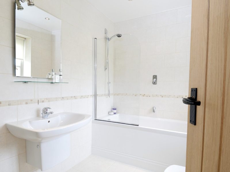 Baño blanco reformado con bañera, mampara de vidrio, lavabo de pedestal y cenefa de madera